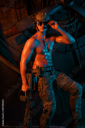 A young muscular soldier in a helmet with a naked torso on an urban background in orange and blue light.