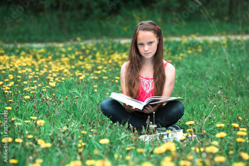 Teen girl reading a book in a green field and looking at the camera.