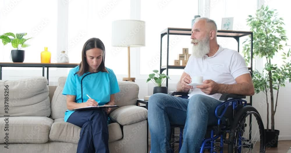 Female healthcare assistant taking notes on health of disabled old man drinking coffee on wheelchair at home Female healthcare assistant taking notes on health of disabled old man drinking coffee on wheelchair at home