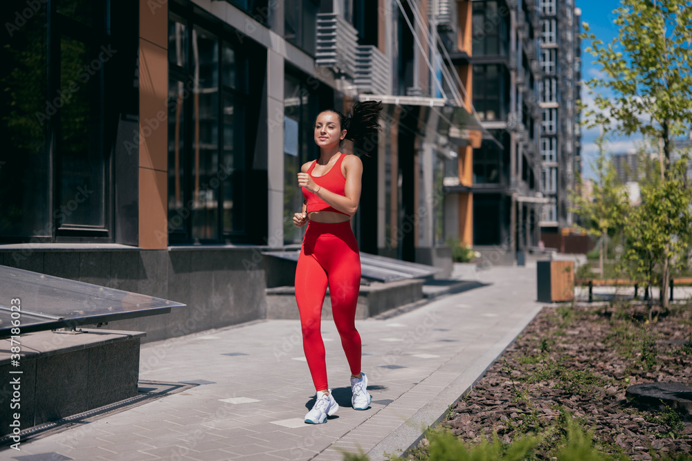 Sporty pretty woman jogging on tiled pavement in urban area. Fit girl with dark ponytail wearing in red sport top, leggins and white sheakers. Apartment block and trees in background.