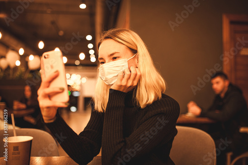 Young woman sits in a cafe, quarantine time. Happy blonde is resting in a cafe, wearing a mask and talking on the phone. Portrait photo of a woman in a black sweater 