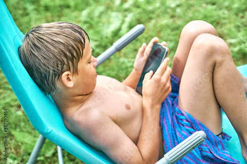 Gadget addiction. Wet boy sitting on sun lounger and playing on smartphone photo