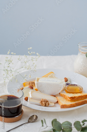 Various types of cheese with nuts and honey in a white plate on a light table and a mug of black coffee. dinner at restaurant