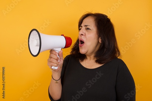 Middle aged Arab woman wearing casual black sweater standing against yellow background, speaking through Megaphone with Available Copy Space. Politics, rights, information. 