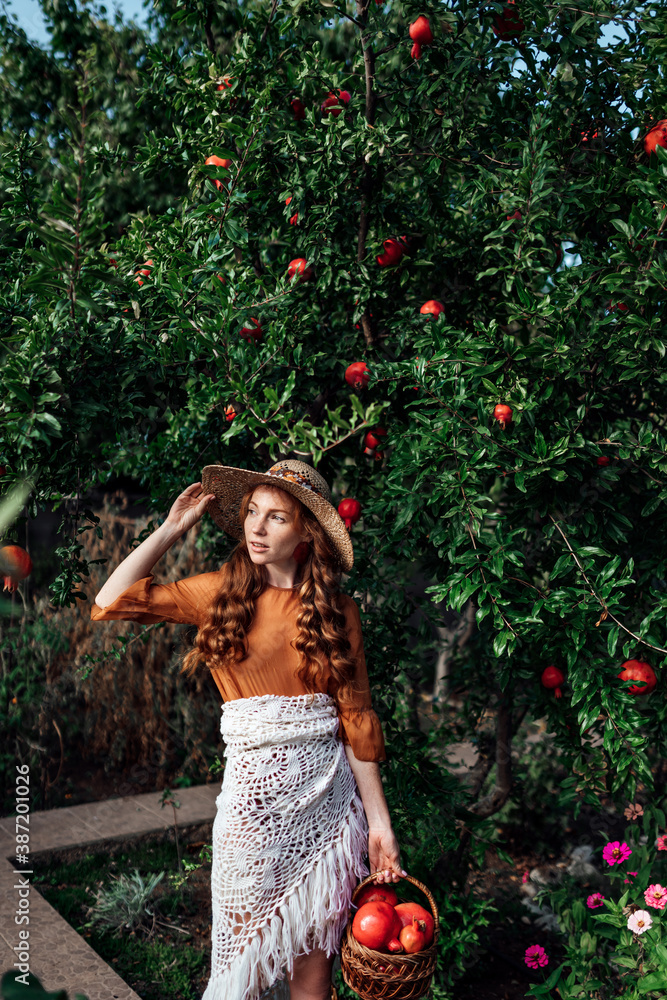 Red pomegranate garden with a happy redhead girl in a hat in an orange dress and white shawl. Woman with big red fruits in .fruit basket near green tree