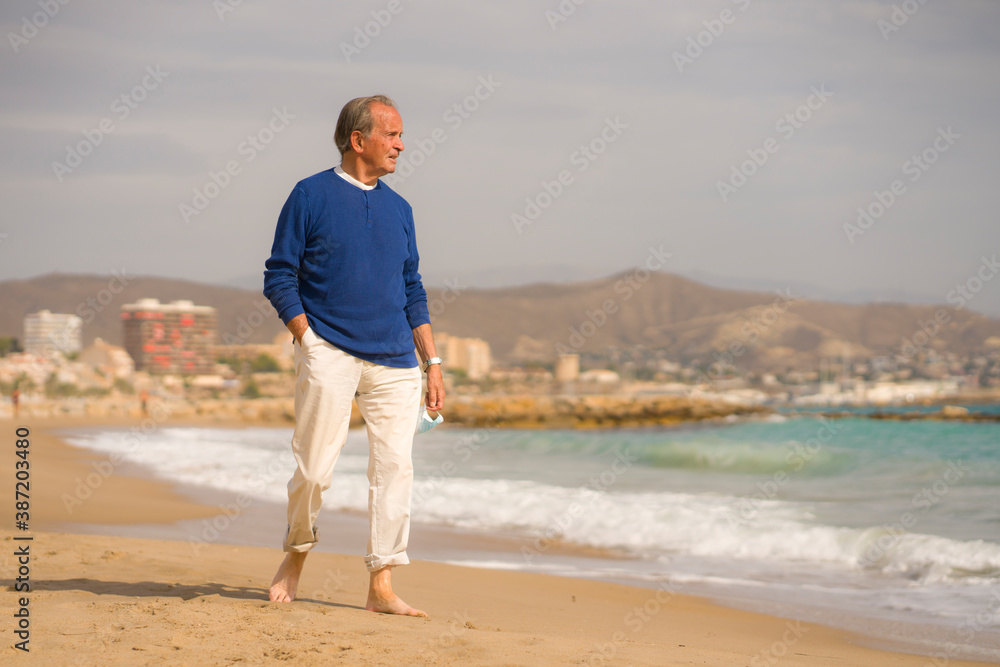 Senior pensioner sitting relaxed on the beach - retired old man on his 70s looking at the sea thoughtful and contemplative with surgical mask on his hand