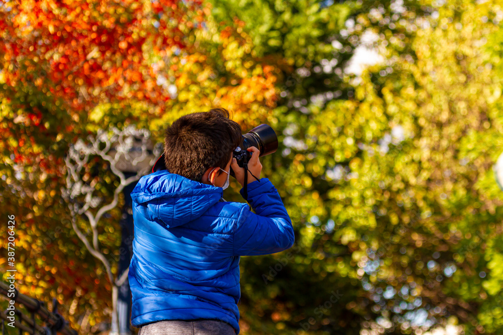 A caucasian boy wearing face mask due to COVID-19 pandemic is taking a photo of the autumn landscape using a professional DSLR camera. Colorful trees in a park are seen in blurred background. A caucasian boy wearing face mask due to COVID-19 pandemic is taking a photo of the autumn landscape using a professional DSLR camera. Colorful trees in a park are seen in blurred background.