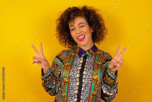 Indoor portrait of young caucasian female isolated over gray background with optimistic smile, showing peace or victory gesture with both hands, looking friendly. V sign.