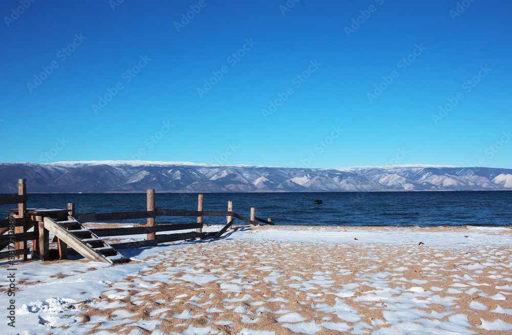 The snow-covered autumn beach of Lake Baikal, with mountains in the background. Staircase over the fence of a private area