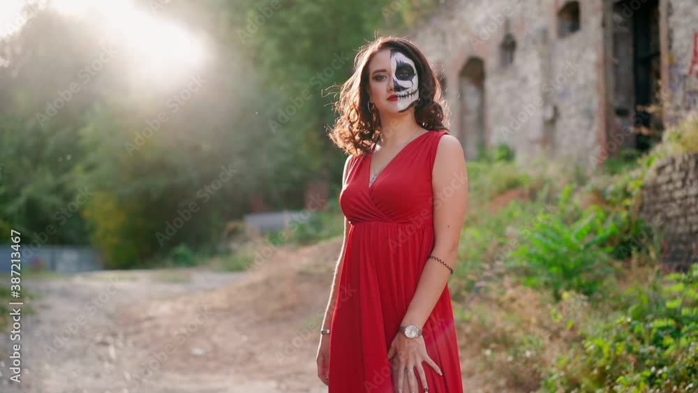 Portrait beautiful slim woman in a red evening dress and awesome Halloween makeup stands against backdrop an abandoned building into sunset light. Greasepaint for Holiday on October 31. Slow motion. Portrait beautiful slim woman in a red evening dress and awesome Halloween makeup stands against backdrop an abandoned building into sunset light. Greasepaint for Holiday on October 31. Slow motion.