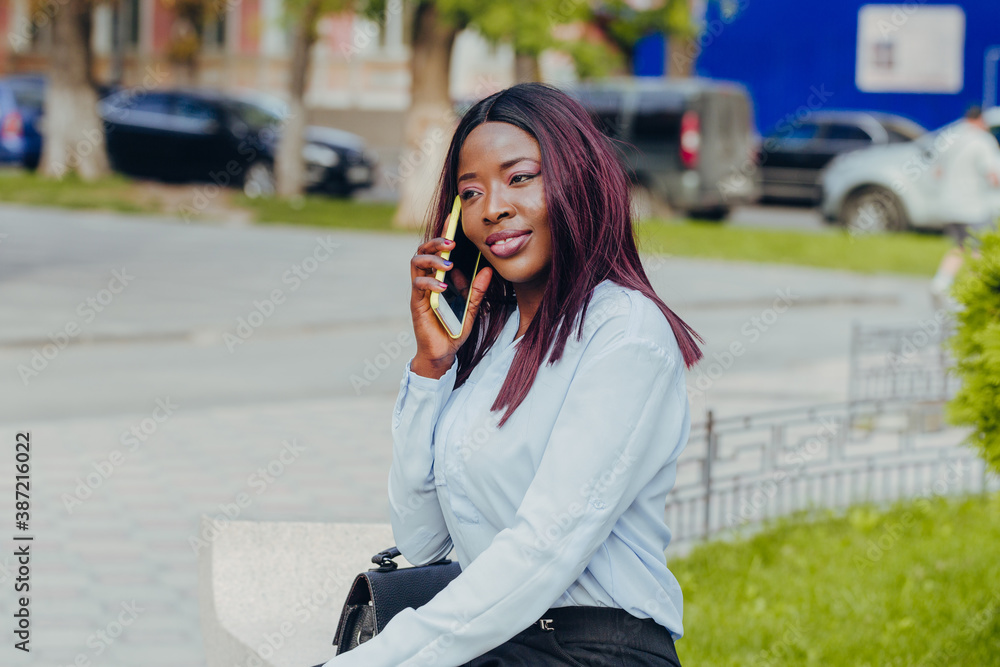 smiling young african american girl with pink hair and black hat sitting on a bench with a mobile phone outdoors on a sunny day. smiling young african american girl with pink hair and black hat sitting on a bench with a mobile phone outdoors on a sunny day.