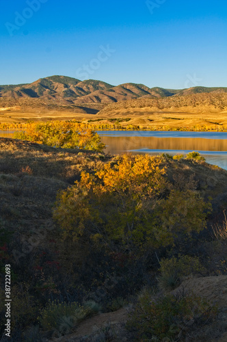 The warm glow of the morning sun bathes the top of autumn trees and the distant mountains on a chilly October morning at Chatfield Lake in Colorado.