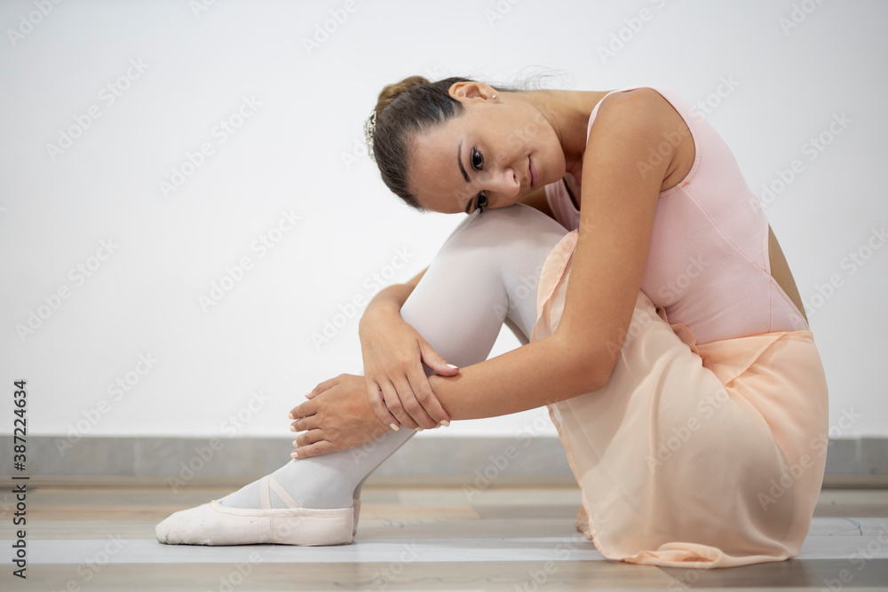 Beautiful professional ballerina in pink dress sitting and stretching her body on wooden floor in ballet studio