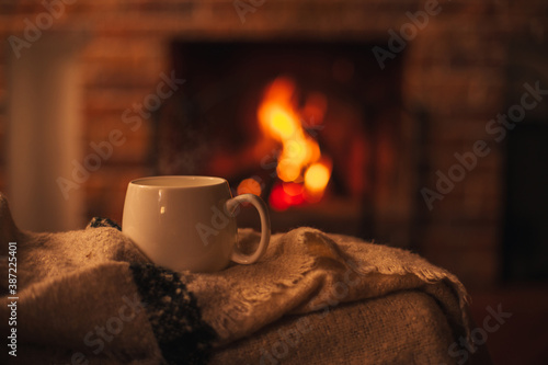 Mug with hot tea standing on a chair with woolen blanket in a cozy living room with fireplace.