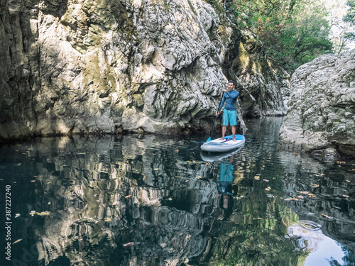 Young man standing on SUP with paddle and floating on river water in rocky canyon