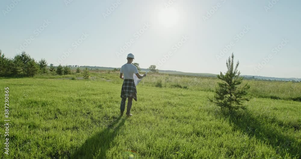 Male architect reading draft in field. Pan around view of man in hardhat unrolling and examining blueprint while standing in green grassy meadow in countryside Male architect reading draft in field. Pan around view of man in hardhat unrolling and examining blueprint while standing in green grassy meadow in countryside