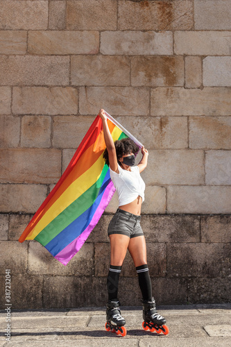Beautiful African American woman wears a face mask while skating with the lgbtq flag of gay pride in the street on a sunny day
