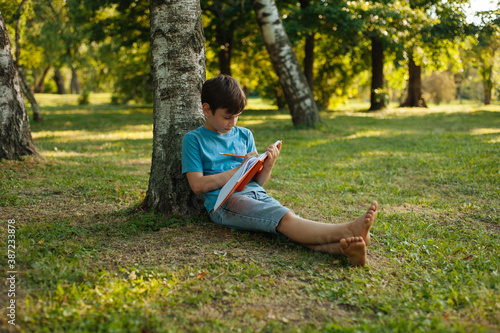 A kid leaning on a tree in park is holding his journal and writing in it photo