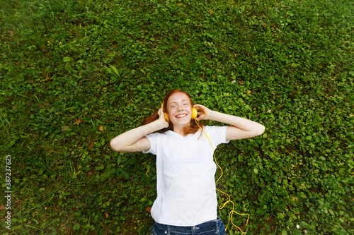 red-haired girl listens to lessons with headphones. Lying on the grass