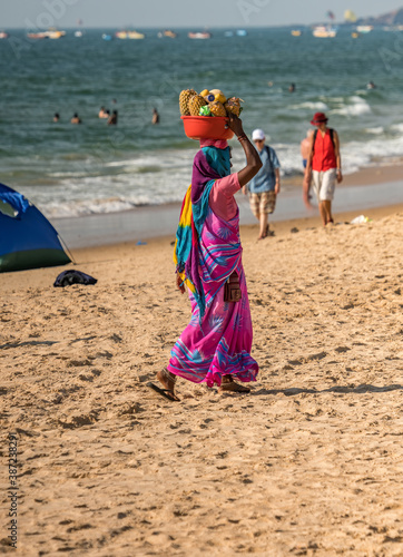 Unidentified Indian woman in colorful pink blue sari dress carrying fresh fruits in red basin at her head for selling at shoreline to tourists. Sunny day at beach in India. Side view