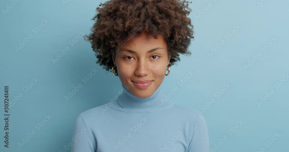 Lovely young lady with Afro hair looks happily at camera smiles broadly winks eyes expresses positive emotions has glad expression hears something pleasant enjoys posing for photo isolated on blue Lovely young lady with Afro hair looks happily at camera smiles broadly winks eyes expresses positive emotions has glad expression hears something pleasant enjoys posing for photo isolated on blue