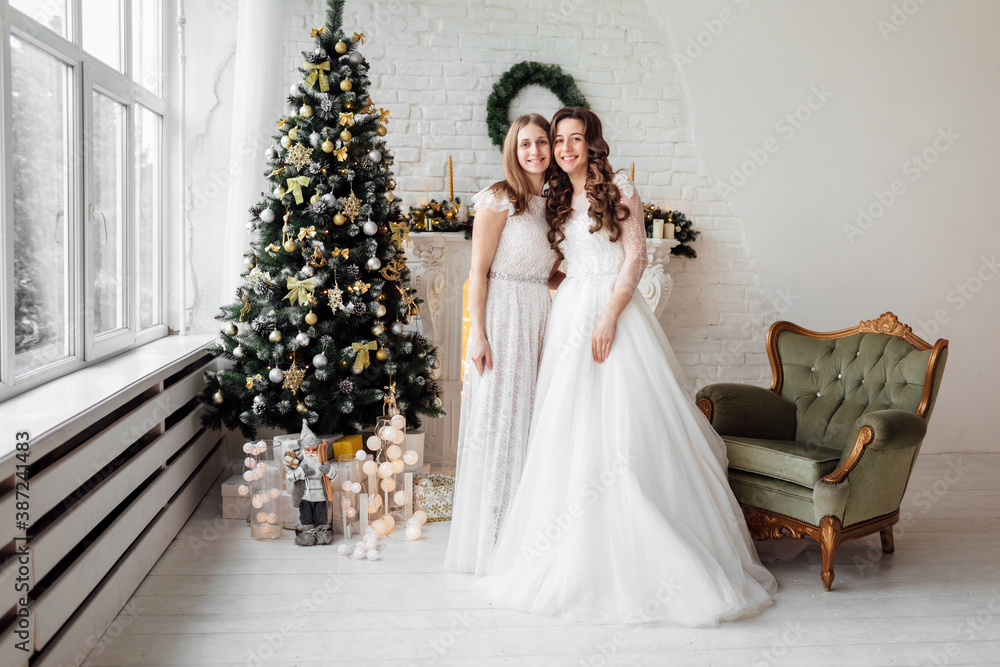 Fototapeta premium Beautiful bride in luxury wedding dress and pretty bridesmaid in dresse in decorated christmas studio. weddind in Christmas