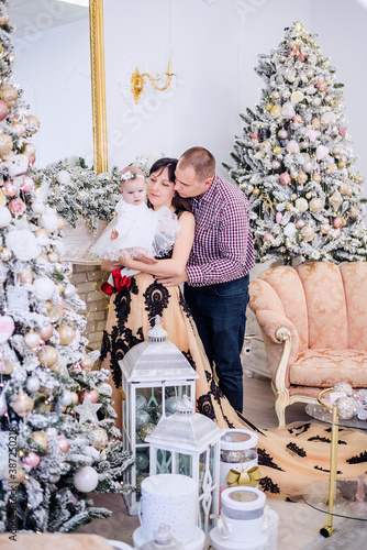 A young family is festively dressed for the evening, holding a little baby in their arms against the background of snow-covered Christmas trees with garlands of lights, gifts, a white fireplace. Card © farmuty