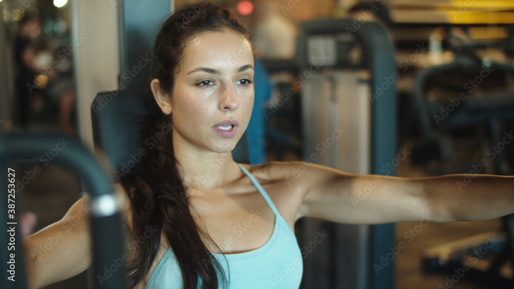 Obraz premium Sporty girl exercising on simulator in club. Fit girl making bicep curls at gym