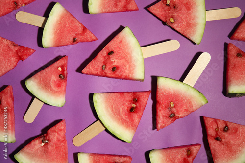 Watermelon slices with wooden sticks on purple background, flat lay