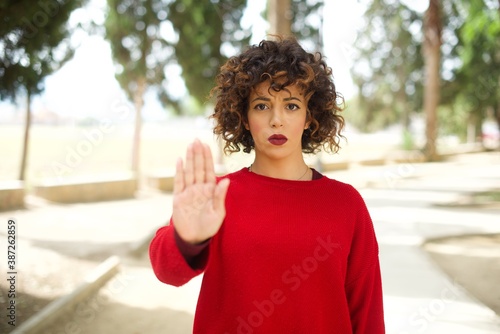 Young arab woman wearing casual red sweater in the street shows stop sign prohibition symbol keeps palm forward to camera with strict expression © Roquillo
