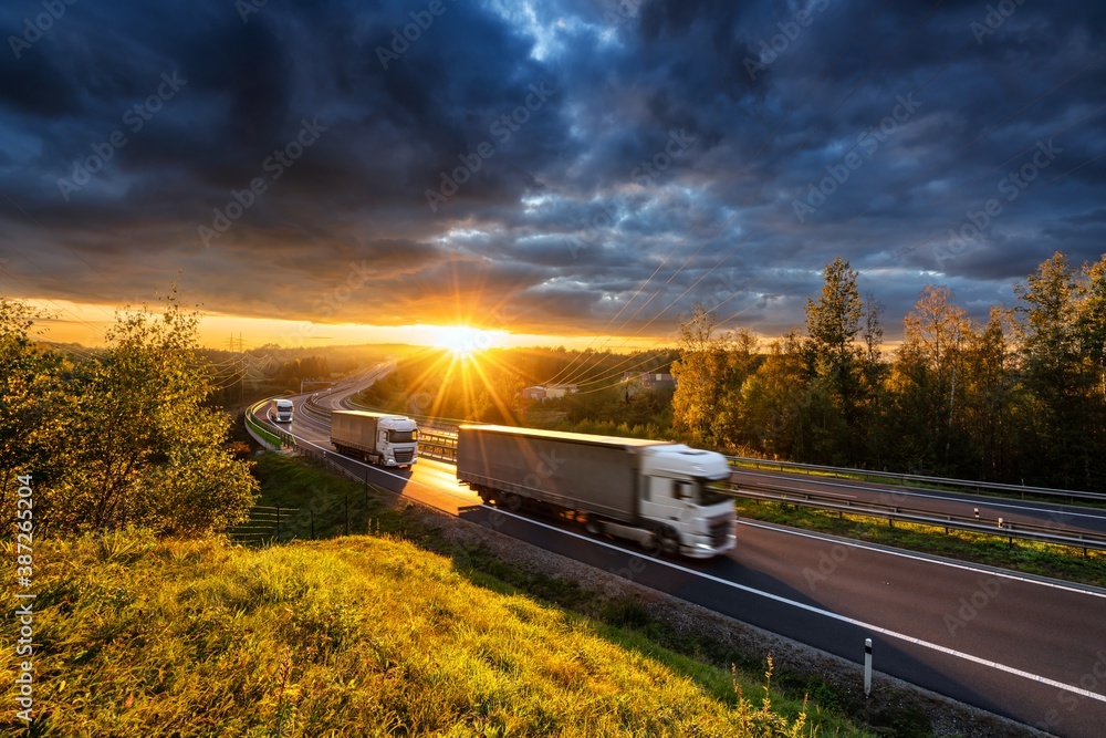 Three motion blurred trucks driving on the asphalt highway in forested landscape in the golden rays of the sunset with dark cloud - obrazy, fototapety, plakaty 