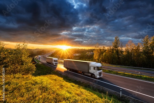 Obraz na płótnie Three motion blurred trucks driving on the asphalt highway in forested landscape in the golden rays of the sunset with dark cloud w salonie