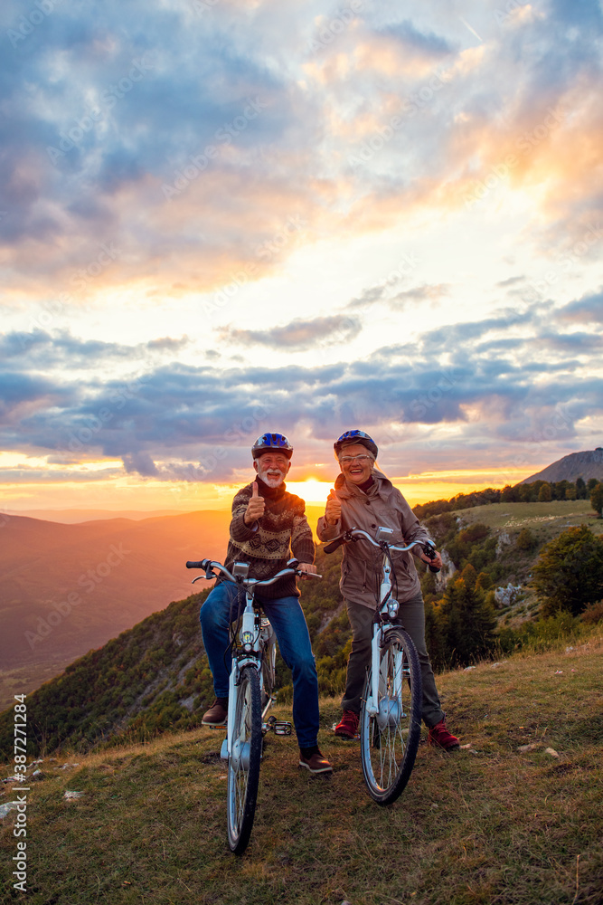 Fototapeta premium Elderly man and woman on e-bikes holding their thumbs up on mountain. Beautiful sunset at background.