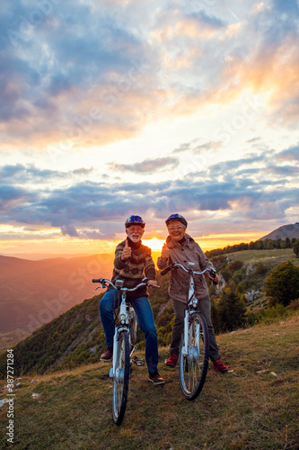 Elderly man and woman on e-bikes holding their thumbs up on mountain. Beautiful sunset at background.