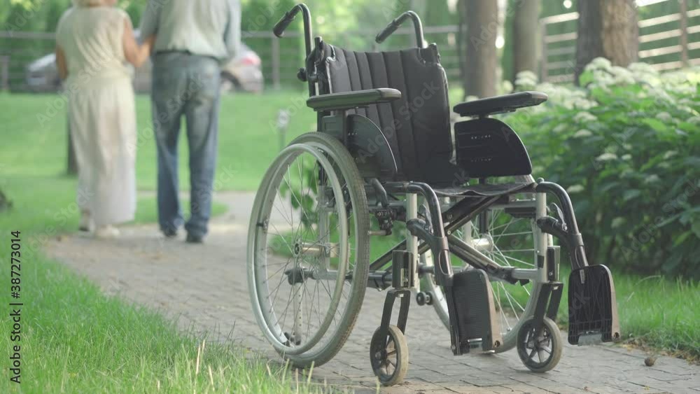 Wheelchair standing on park alley in sunlight with blurred senior Caucasian couple walking away at the background. Old husband and wife strolling outdoors with weak spouse on summer day. Wheelchair standing on park alley in sunlight with blurred senior Caucasian couple walking away at the background. Old husband and wife strolling outdoors with weak spouse on summer day.