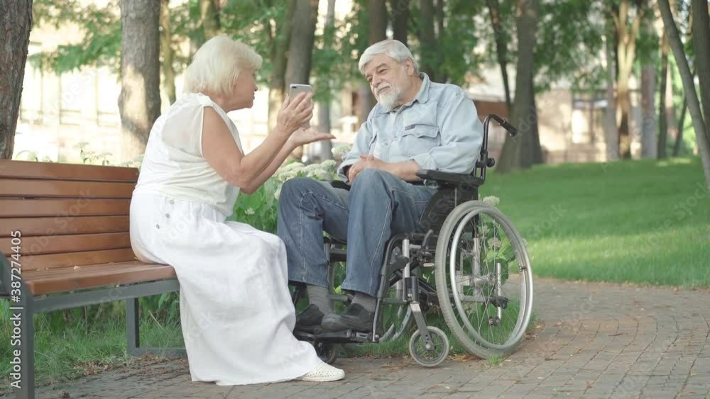 Wide shot of positive smiling husband and wife using smartphone in sunny park. Loving senior Caucasian woman sitting on bench with disabled husband in wheelchair and talking. Wide shot of positive smiling husband and wife using smartphone in sunny park. Loving senior Caucasian woman sitting on bench with disabled husband in wheelchair and talking.