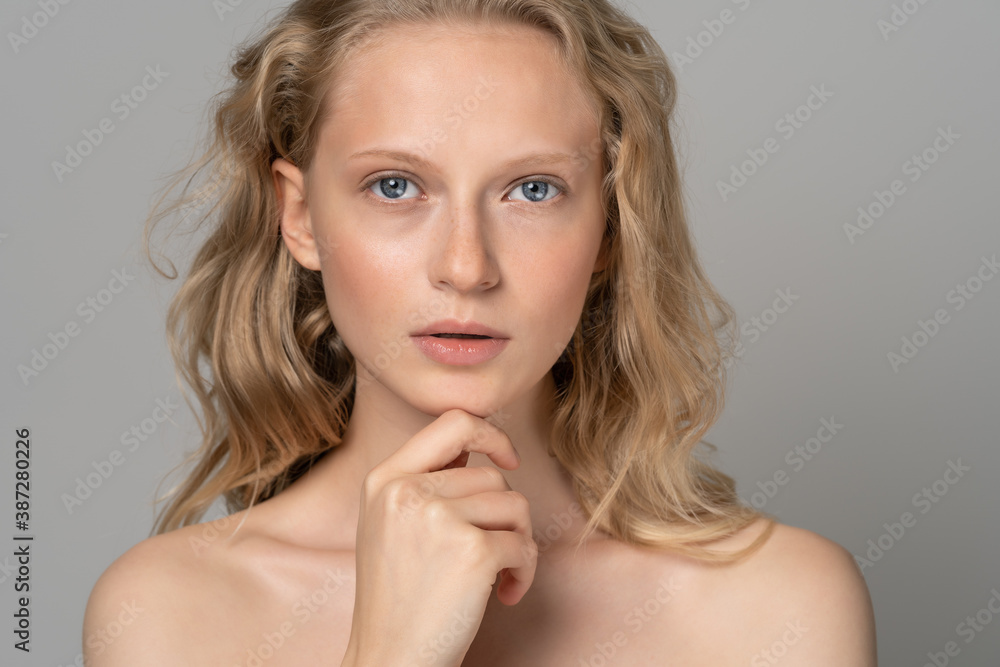 Close up of beautiful young woman face with blue eyes, curly natural blonde hair, has no makeup, touching her chin, standing shirtless with bare shoulders, looking at camera. Studio grey background. Close up of beautiful young woman face with blue eyes, curly natural blonde hair, has no makeup, touching her chin, standing shirtless with bare shoulders, looking at camera. Studio grey background.