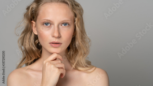 Close up of beautiful young woman face with blue eyes, curly natural blonde hair, has no makeup, touching her chin, standing shirtless with bare shoulders, looking at camera. Studio grey background.