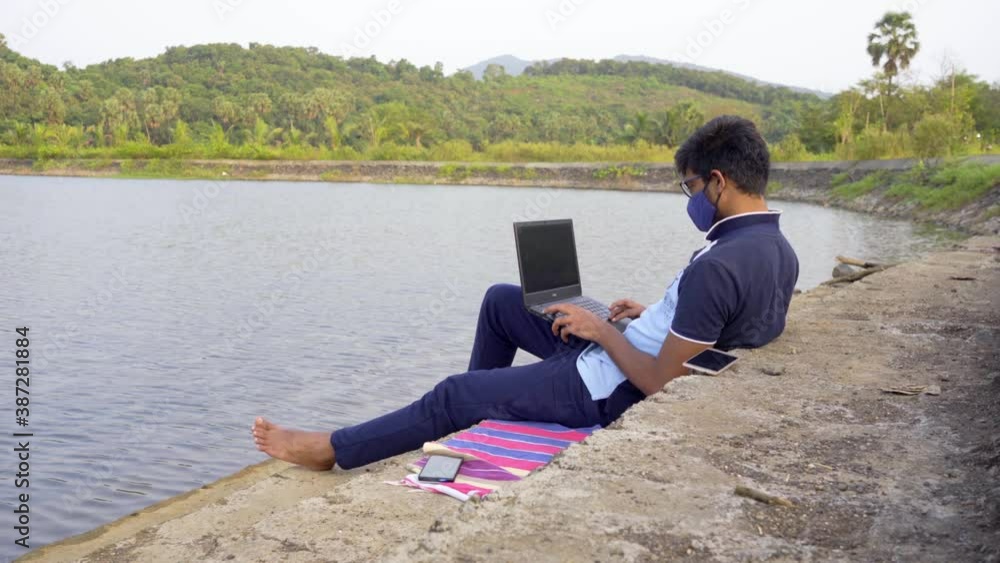 working remotely from laptop waring mask location lakeside India new normal wide shot relaxed working remotely from laptop waring mask location lakeside India new normal wide shot relaxed