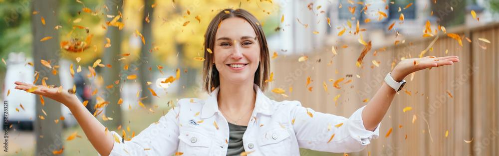 Happy middle age Caucasian woman throwing autumn fall yellow leaves. Young woman with short haircut enjoying life in park outdoors. Little memorable moments of life. Web banner header. - obrazy, fototapety, plakaty 