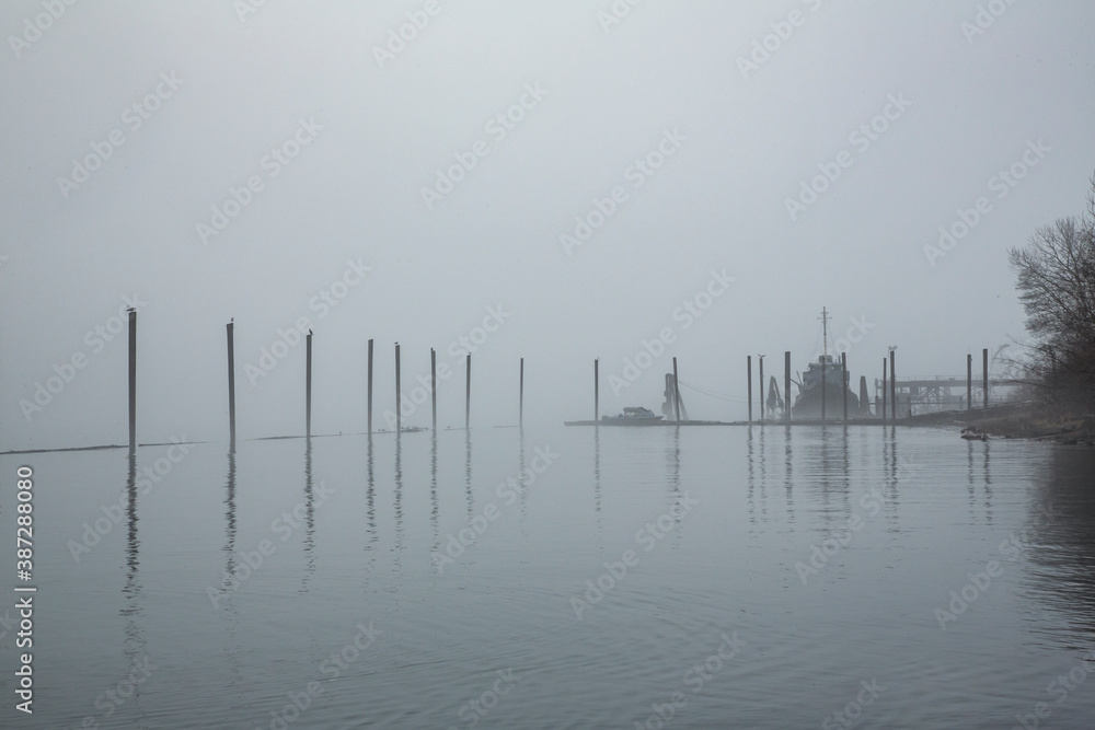 A foggy view of river pylons with the silhouette  of a small boat
