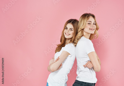 Two girlfriends in white T-shirts stand with their backs to each other on a pink background