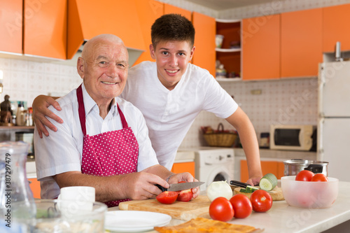 Cheerful teenage boy hugging his smiling elderly grandfather, posing together in comfy home kitchen