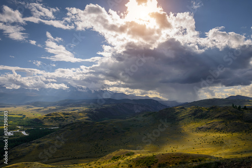 Majestic panorama of mountain plain on the background of snow-covered ridge before thunderstorm. Sun's rays break through huge clouds and beautifully illuminate green steppe and winding river.