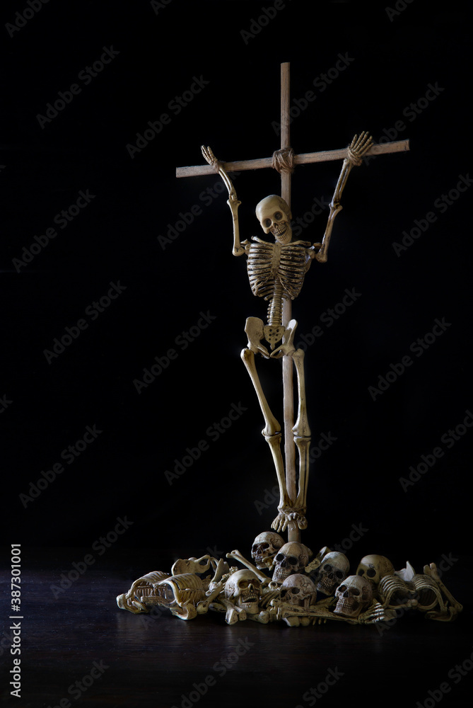 Skeleton tied up with crucifix wich has pile of skulls and bone below all put on dark background