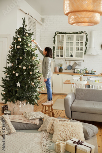 Little girl standing on the chair and decorating Christmas tree alone for Christmas Day in the living room at home