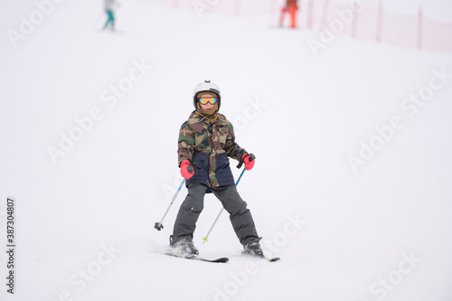 active little sport boy in helmet and khaki ski suit riding skiing with poles on winter white snow slope