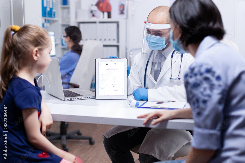 Pediatrician showing skeleton on tablet pc to mother and daugther wearing face mask agasint coronavirus during consultation. Doctor specialist providing health care service radiographic treatment Pediatrician showing skeleton on tablet pc to mother and daugther wearing face mask agasint coronavirus during consultation. Doctor specialist providing health care service radiographic treatment