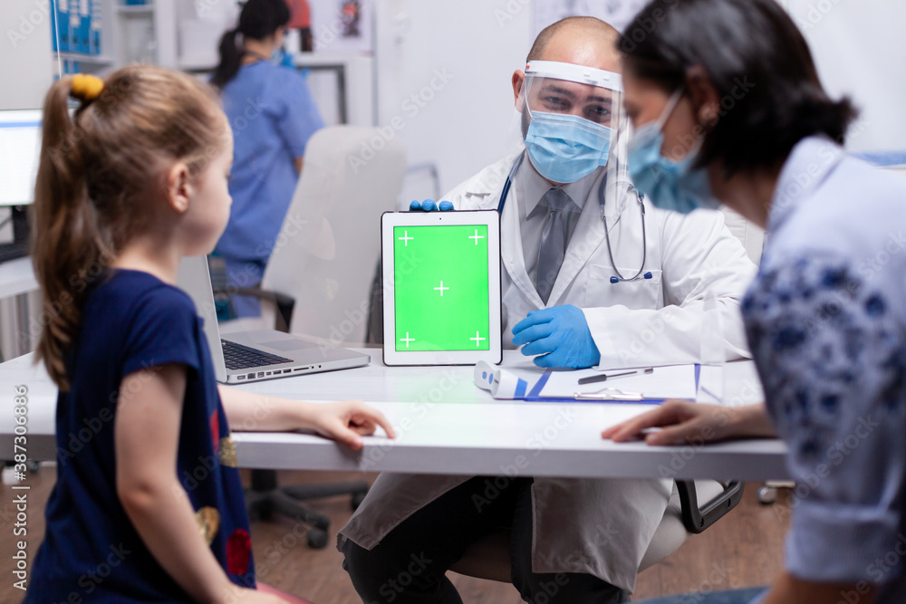 Doctor with face mask against coronavirus using green screen tablet in medical office. Pediatrician doctor in protection gloves and mask providing clinic medicinal health care services.