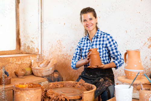 Portrait of smiling girl sitting behind pottery wheel and demonstrating finished ceramic jug